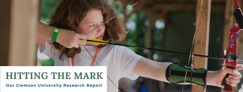 A Kudzu camper draws her bow and aims an arrow at an offscreen target. A title is overlaid on the lower left corner that says "Hitting the Mark: Our Clemson University Research Report."